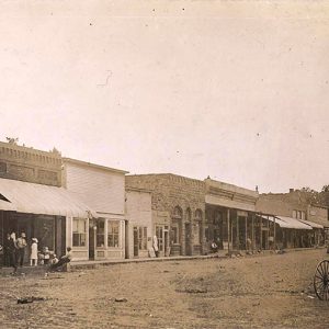 People on sidewalk outside single and multistory buildings on dirt street with horse drawn carriage in the foreground