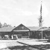 Faded black and white lodge building with three cars and flag pole in front yard and person standing by one car