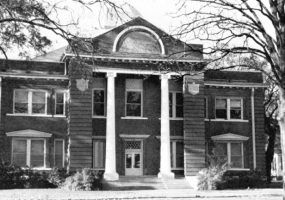 Two-story brick building with arched attic window and two columns with trees and grounds