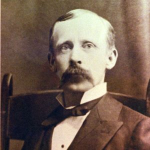 White man in suit with large mustache sitting in wooden chair