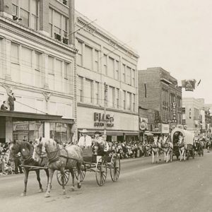 Horse-drawn wagons in parade on city street with crowds of people standing in front of multistory buildings