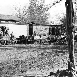 Men and horse-drawn wagons outside wall with building behind it