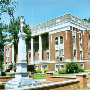 Monument on pedestal with statue on top in front of multistory brick building with white columns