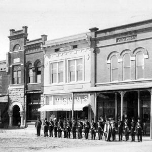 Group of men in uniform standing in line in front of row of brick store fronts on dirt road