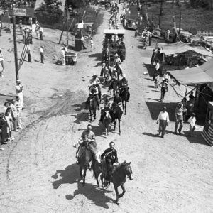 Parade of people on horses with spectators watching on a dirt road