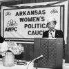 White woman and African-American woman speaking at lecterns on table with "Arkansas Women's Political Caucus" banner on the wall behind them