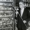 Older white man with glasses in suit standing before shelves filled with various items