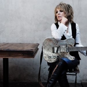 White woman with black and white outfit and heavy eye makeup sitting at a wooden table holding a silver Fender Telecaster guitar