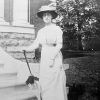 White woman in hat and dress leaning on closed parasol outside brick building