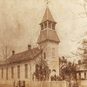 Wood frame church with steeple men on front steps women on sidewalk picket fence winter