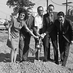 White men in suits with white woman in dress and sunglasses posing with hands on a shovel in dirt