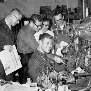 Group of white men with linotype machine, some holding issues of a newspaper