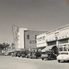 Row of parked cars on street outside brick storefronts and "Joy" movie theater