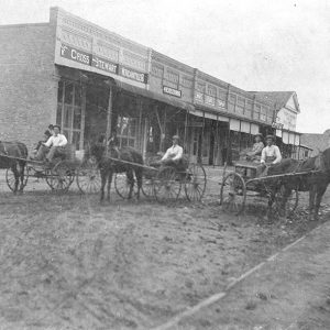 Men in three horse drawn buggies on dirt road with storefronts on left side