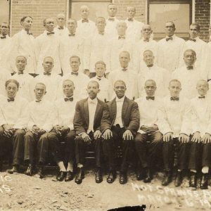 Group of African-American men in matching uniforms with white jackets and bow ties and two in front with matching black suit jackets posing in front of brick building