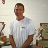 White man smiling in white shirt with Arkansas shaped trophy on table next to him