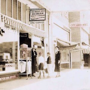 White women and children standing outside ice cream parlor and white men in hats standing outside barbershop behind them