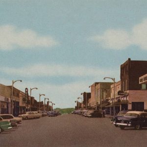 Parked cars on street between rows of multistory brick buildings with signs and street lamps