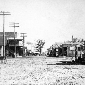 Men pose with wagon on dirt road lined with buildings and telephone poles