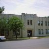 Two-story buff brick building on street with trees and parked cars