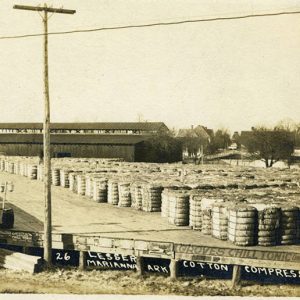 Stacks of cotton bales sitting outside warehouse building with houses in the background