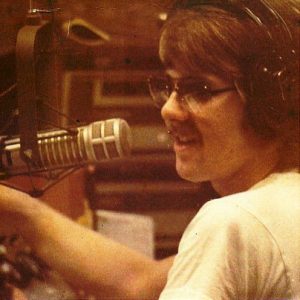 Young white man with mustache and glasses talking into a microphone