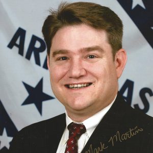 White man in suit and tie smiling with Arkansas flag behind him