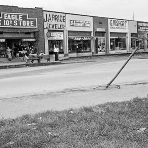 Street and storefronts with signs saying "Eagle 5 and 10 cent store" and "J. A. Price Jeweler" and "G. W. Short General Merchandise" with people on sidewalk and sitting on bench