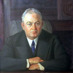 White man in suit sitting at his desk with his arms crossed