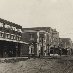 Two-story brick storefronts including one with a sign saying "McKinney Hardware Co." and power lines on dirt road