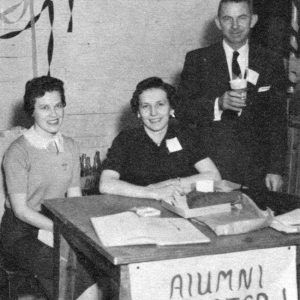 Two white women sitting at table with white man in suit and tie holding a cup standing next to them