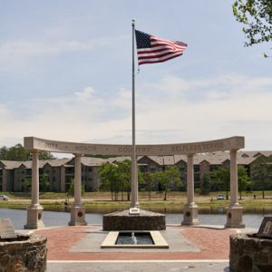 Columns and rounded top with the words "duty honor country selfless service" and flag pole with lake and apartment buildings in the background