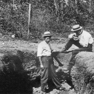 White man in hat standing in pit with another man in a hat sitting on the ground with fence behind them