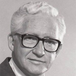 White man with white hair and black-rimmed glasses in suit and tie