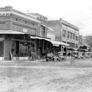 Dirt street with brick buildings and parked cars on the left side