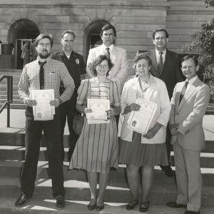 White policeman and three white men in suits posing with white man and women with papers in their hands