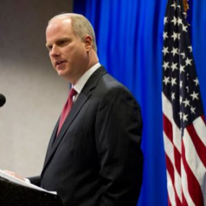 White man in suit speaking at lectern with microphone with American flag behind him and blue curtain