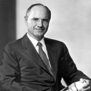 White man in glasses and suit posing in wooden chair with a rolled up newspaper