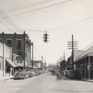 Parked cars on street under traffic lights between rows of buildings with people standing on street corner