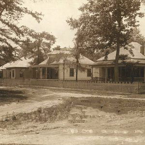 Row of houses with covered porches and trees inside fence on dirt road