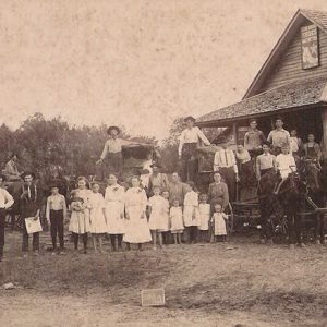 Crowd of white men women and children outside single-story building with covered porch