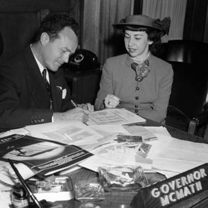 White man in suit signing paper on cluttered desk for white woman in hat and pant suit in his office