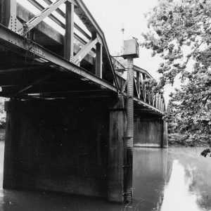 Underside of bridge with steel arch truss and concrete columns in creek