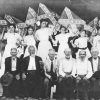 Group of young white women holding Confederate battle flags behind group of older white men