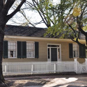 Single-story house with yellow siding and green window shutters behind white picket fence with trees in front yard