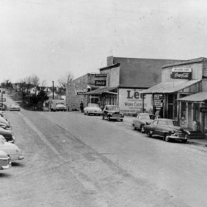 Street with store buildings and parked cars with houses in the distance