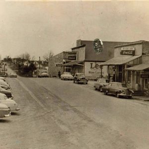 Parked cars on street outside single-story storefronts across from parking lot with line of cars and houses in the background