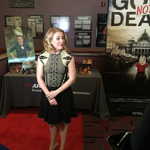 White woman in sleeveless dress standing on red carpet with displays on tables behind her