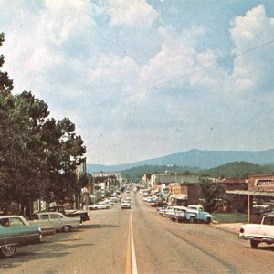 Cars parked outside town buildings on two-lane street with hills in the background