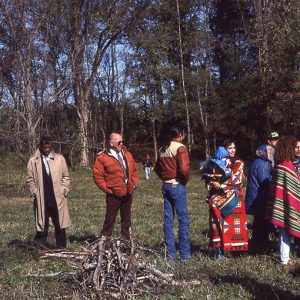 Native American women in traditional clothing and men at mound site with white camera crew and African-American park ranger